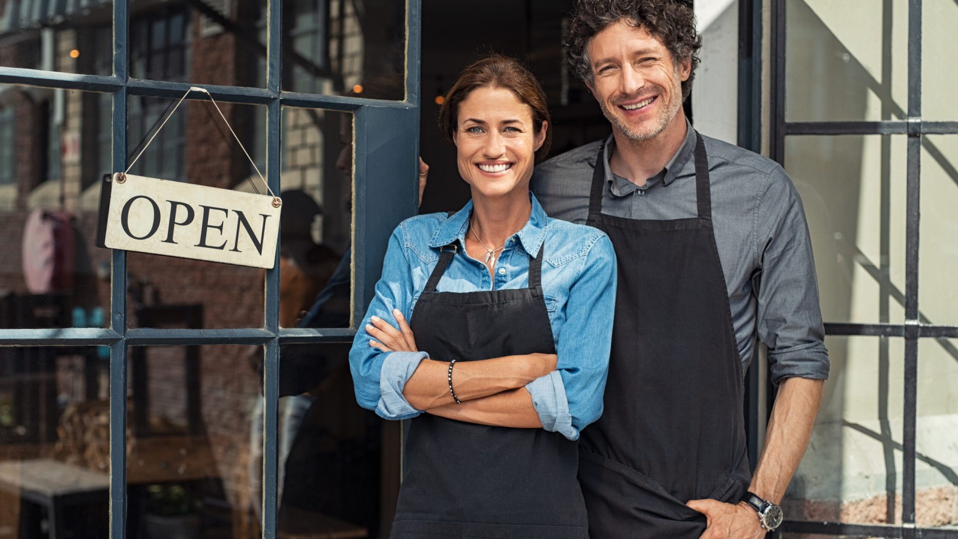 Two cheerful small business owners smiling and looking at camera while standing at entrance door. Happy mature man and mid woman at entrance of newly opened restaurant with open sign board. Smiling couple welcoming customers to small business shop.